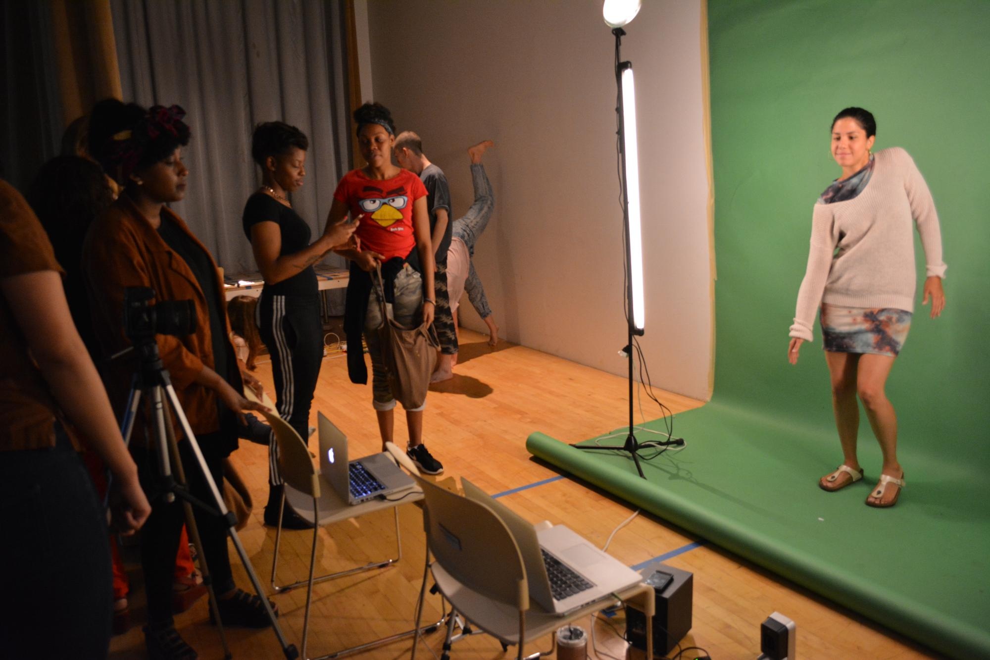 Image of people in a room with media equipment. A person is dancing on a green screen, while others look on.